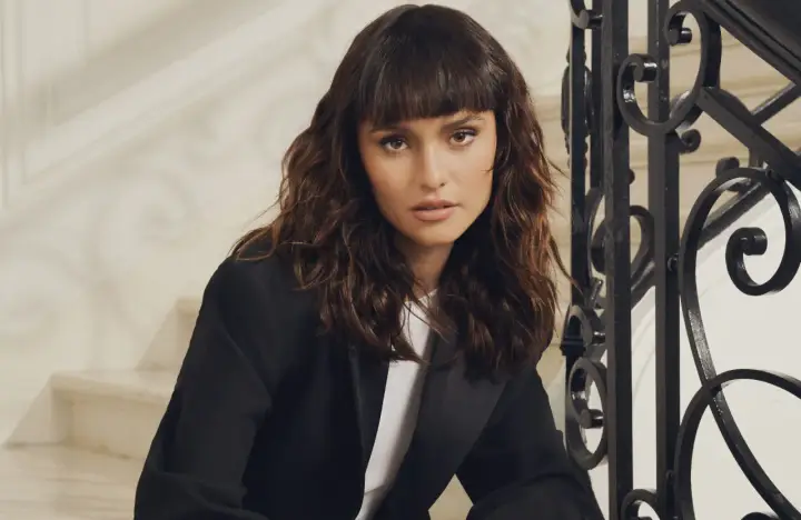 Une femme au regard intense arbore un **wolf cut cheveux bouclés** brun, porté avec un blazer noir.