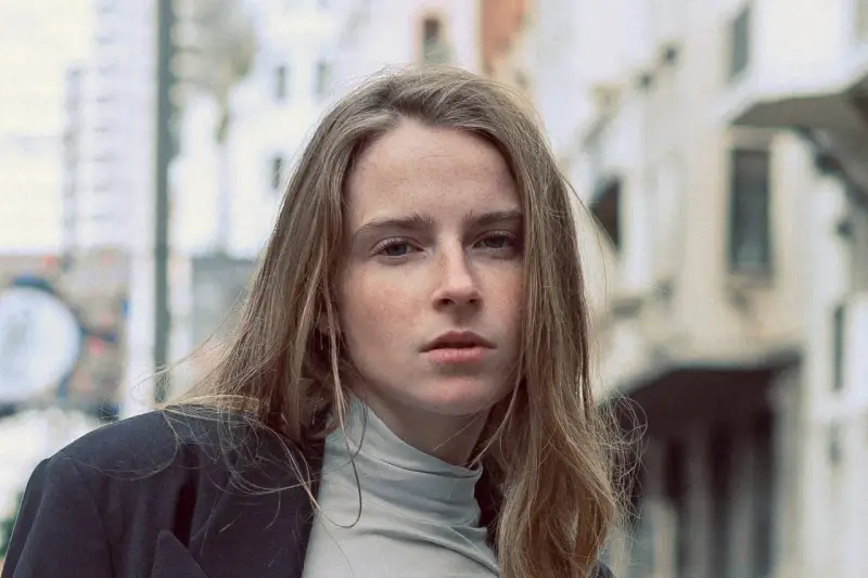 Jeune femme au regard intense, ses cheveux châtain clair cendré ou glacé encadrent son visage parsemé de taches de rousseur. Elle porte un col roulé gris et une veste sombre.